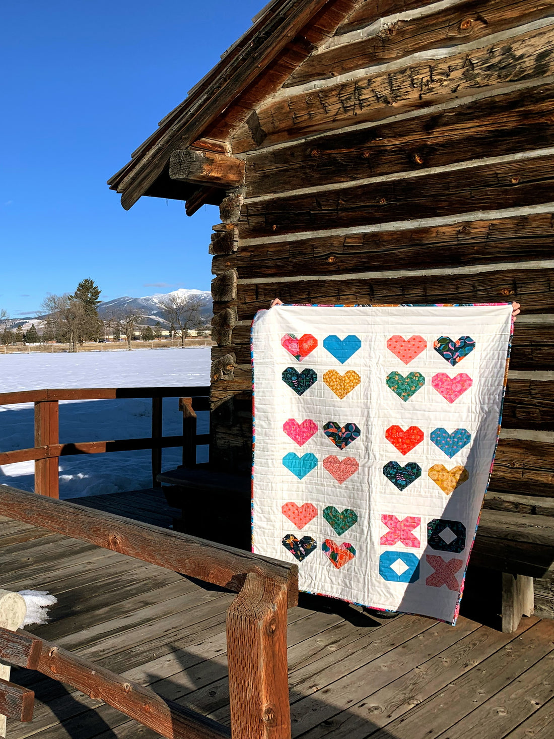 Multi color heart quilt in front of log building with snow and blue sky
