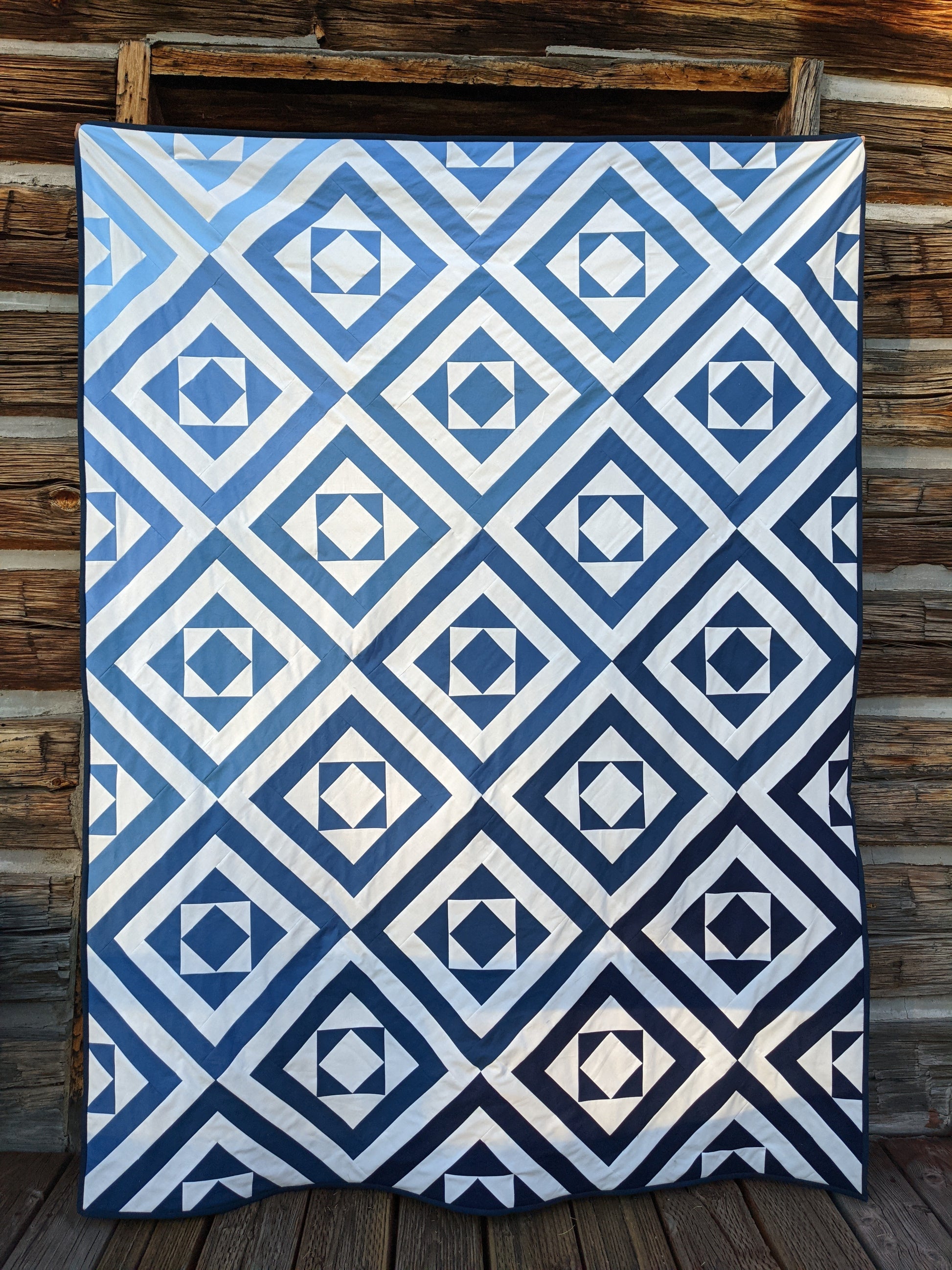 Ombre blue modern quilt square in square in front of log building.